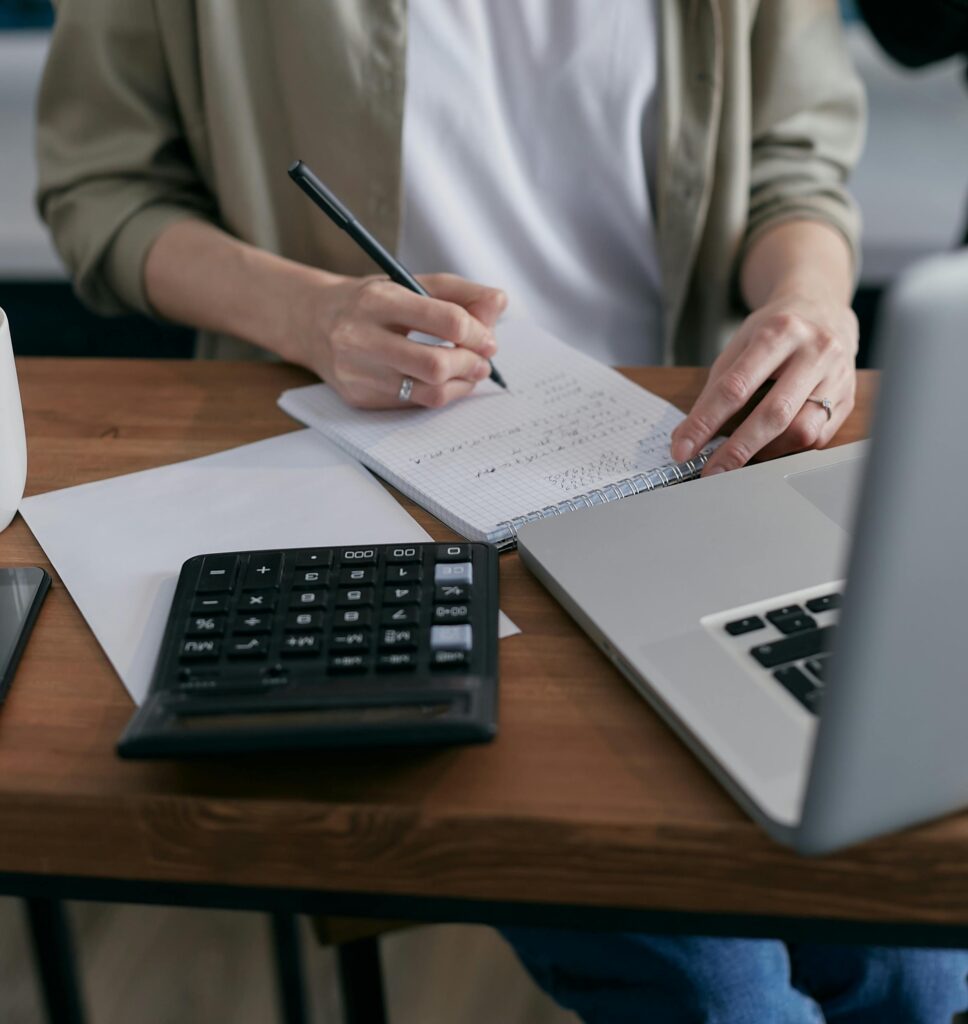 pexels photo 6962993 6962993 A woman writes financial calculations in a notebook, using a calculator and laptop at a wooden desk.